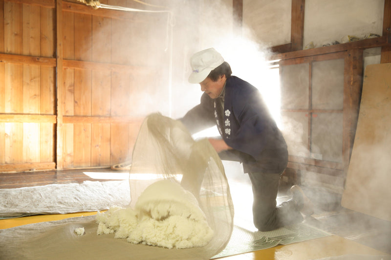 A Dewazakura brewer kneeling in the brewery steam, lifting freshly steamed rice from its cloth.