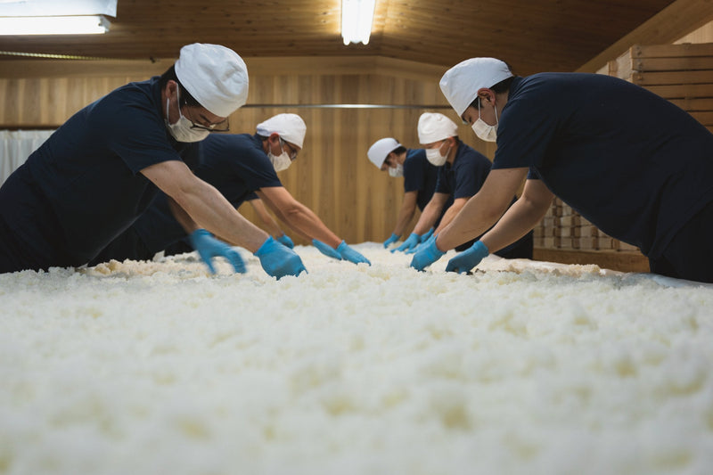 Five people in white caps and blue gloves working rice across a broad table.