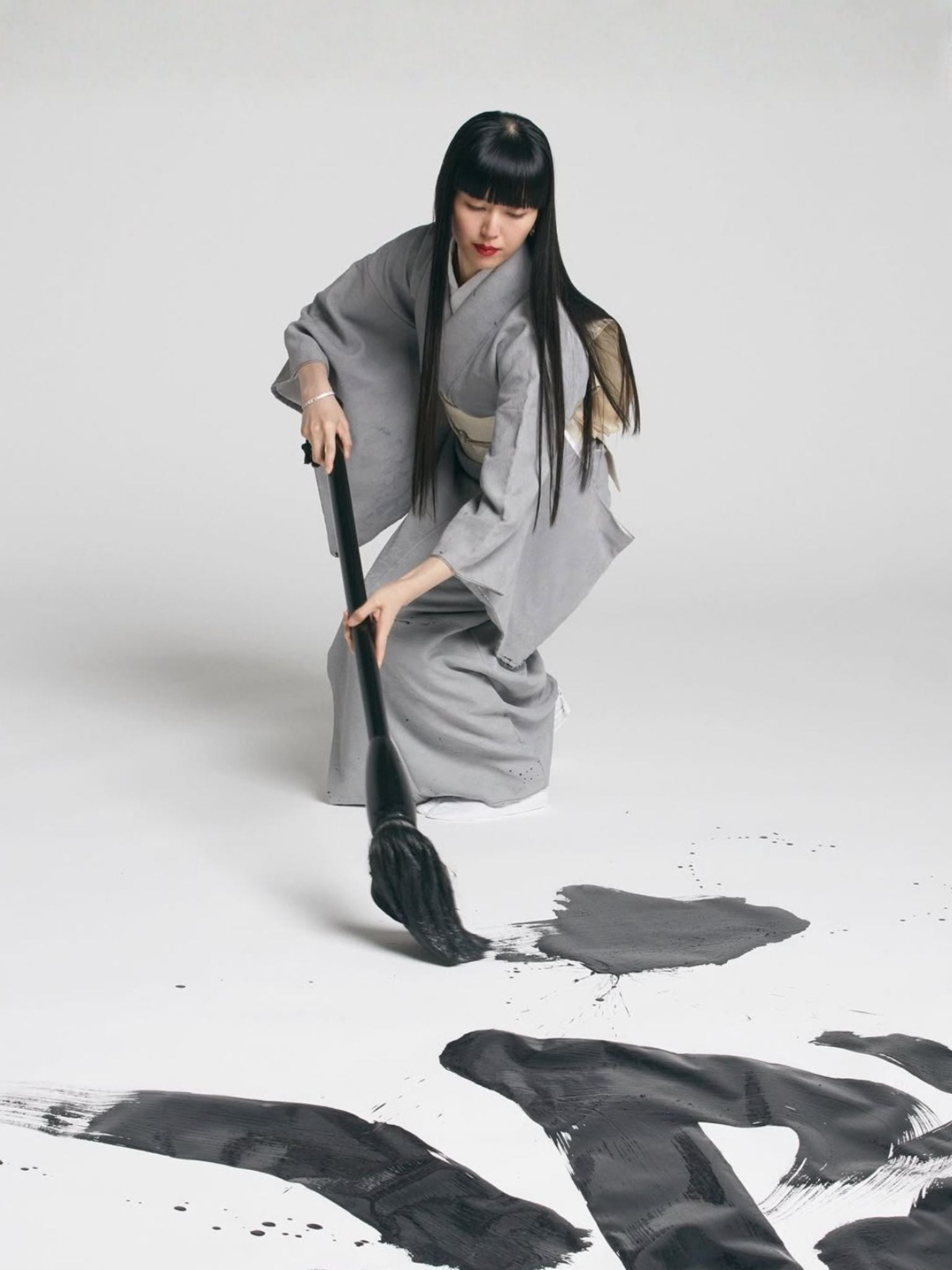 Mamimozi  in traditional attire using a large brush to create black ink splashes on a white background